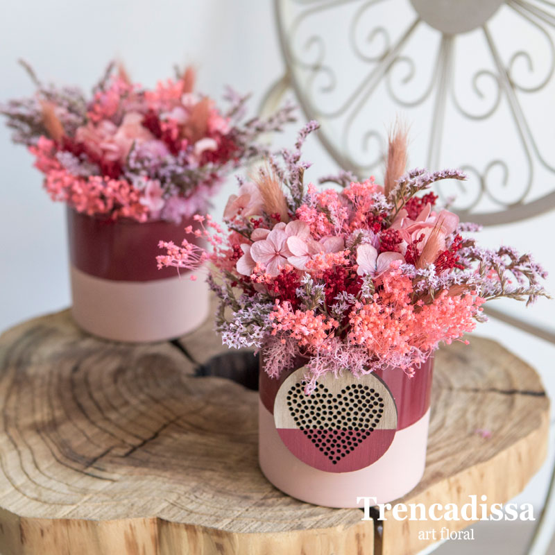 Recipiente de cerámica en tonos rosa y rojo decorado con hortensia preservada y flor seca con corazón decorativo.